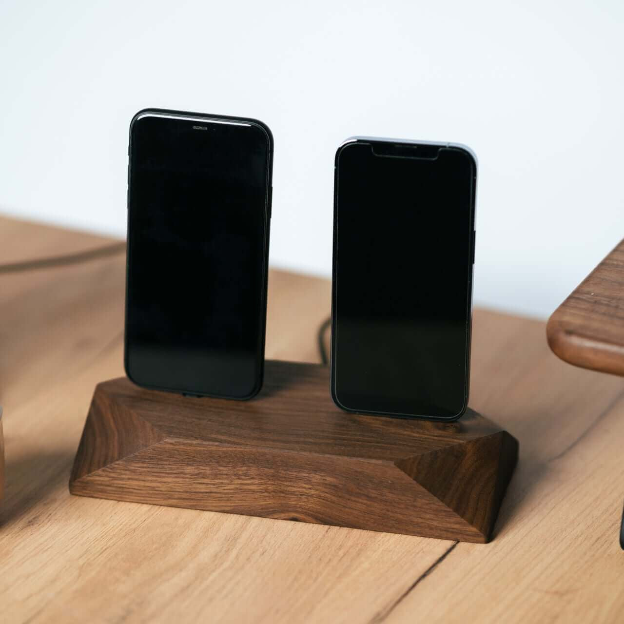 Dual phone dock in stained black oak charging two iPhones simultaneously on a clean desk with headphones nearby.