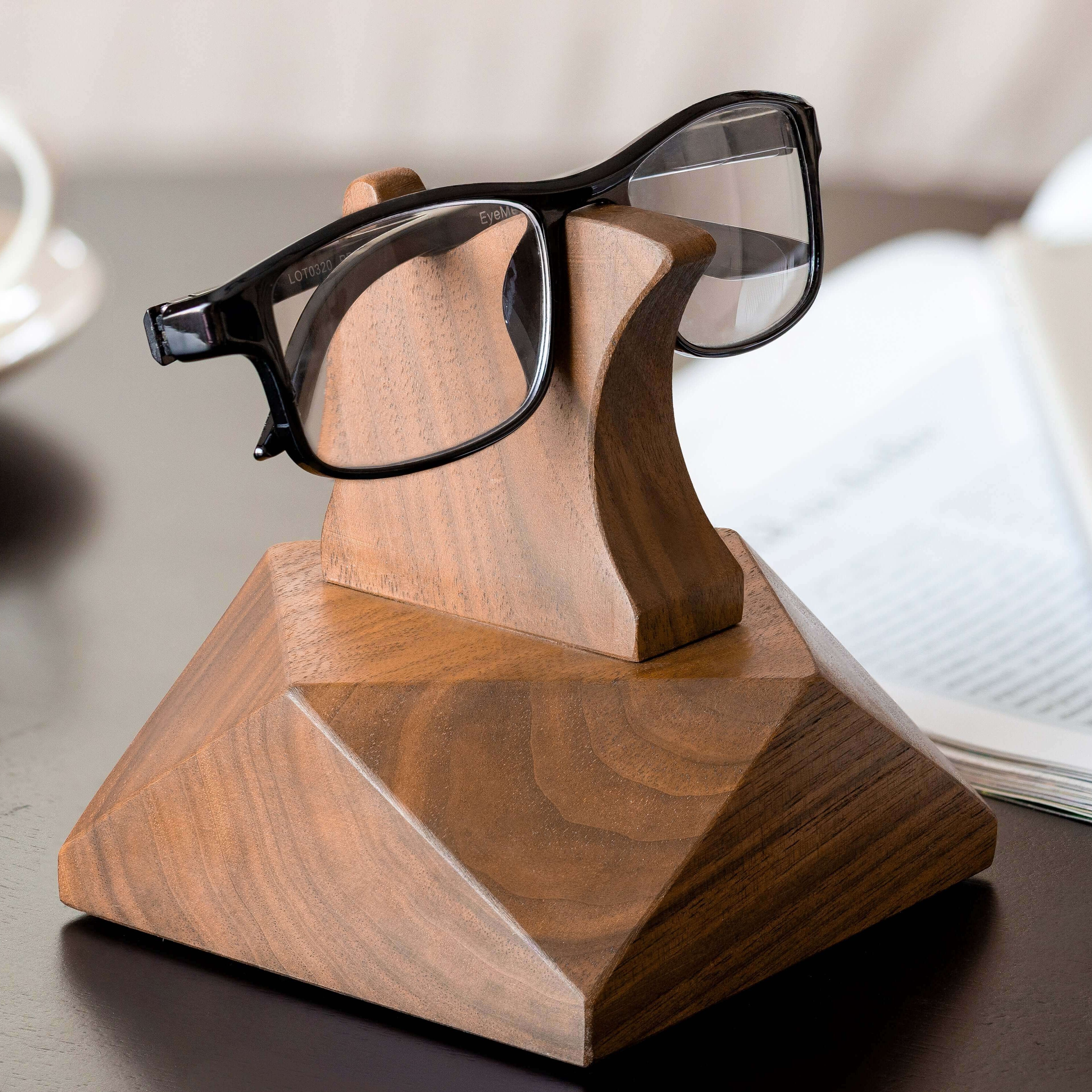 Wooden eyeglasses stand holding black glasses on a desk with a cup and papers in the background.