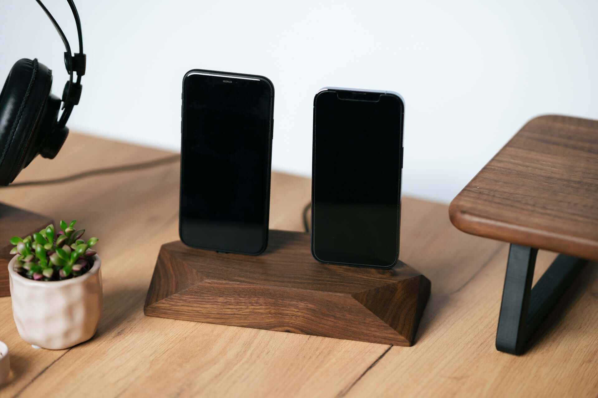 Dual phone dock in stained black oak charging two iPhones simultaneously on a clean desk with headphones nearby.