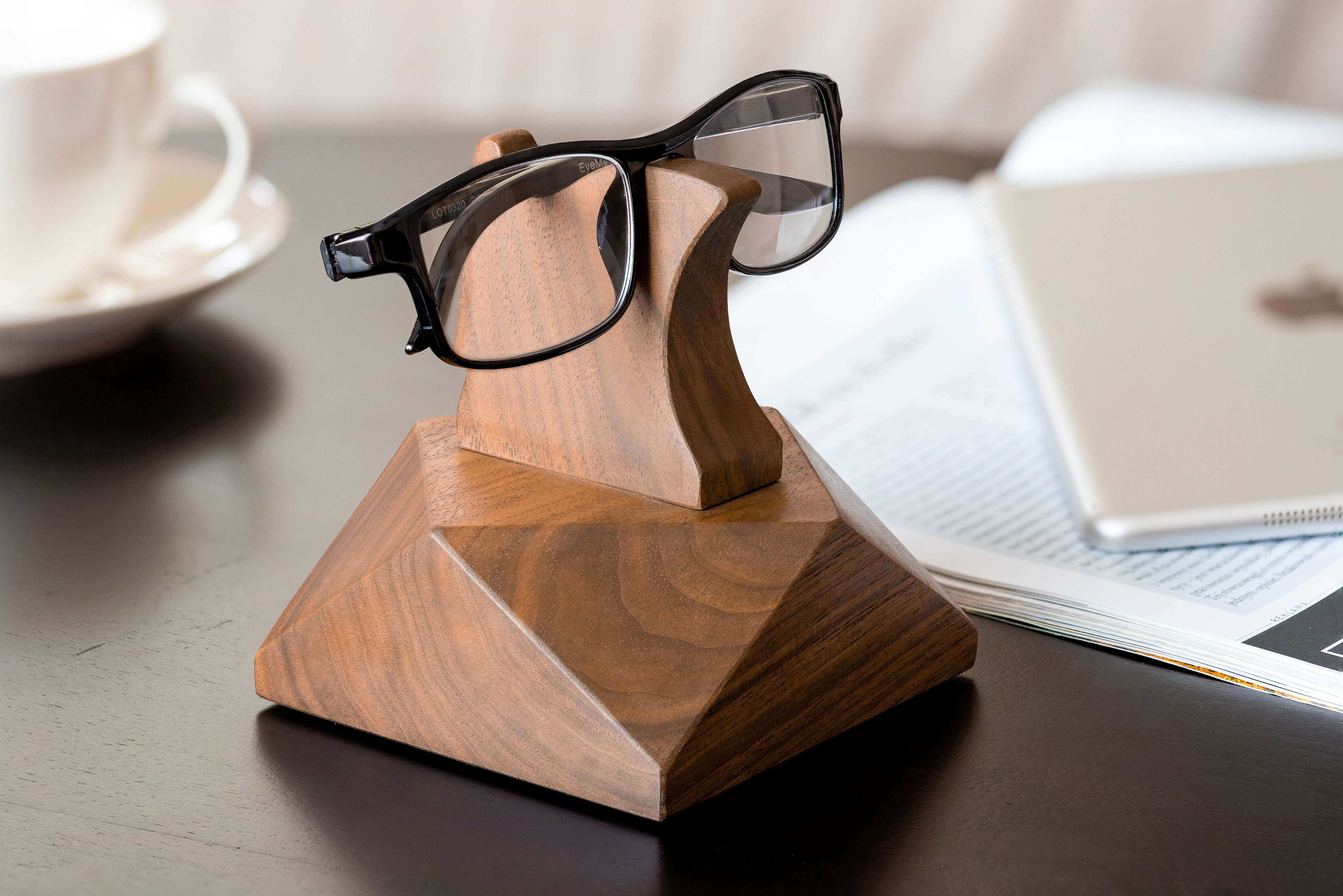 Wooden eyeglasses stand holding black glasses on a desk with a cup and papers in the background.